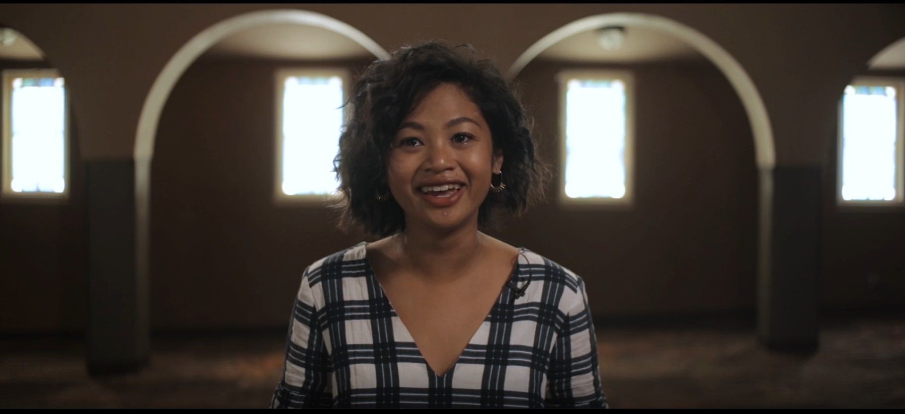 Smiling person standing in a softly lit room with arched windows.